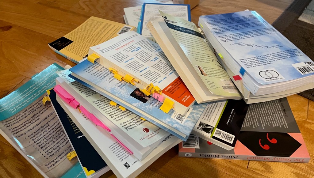 Pile of over 20 books on memoir writing the author has read to learn how to do this. They have many stick tags and notes sticking out of them. 