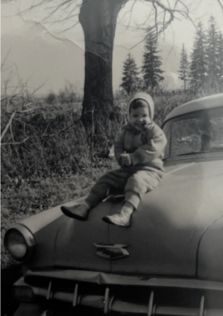 Black-and-white 1957 photo of the author as a 2-year-old toddler in a snowsuit, sitting on the hood of a 1954 Chevy Belair sedan on a sunny late afternoon winter day. Countryside of Torrington CT around Klug Hill Rd.